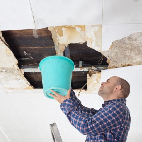 Man Collecting Water In Bucket From Ceiling. Ceiling panels damaged  huge hole in roof from rainwater leakage.Water damaged ceiling .